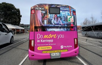 Rear view of a bus stopped in a transport area, covered in a bright pink advertisement reading “Do more you in Karrinyup” with smaller text “Find more to see, do and love” and “Discover Stirling,”